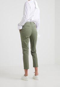 Person i olivgröna kortbyxor, vit skjorta med knappar och vita sneakers, stående med händerna i fickorna mot en enfärgad bakgrund.