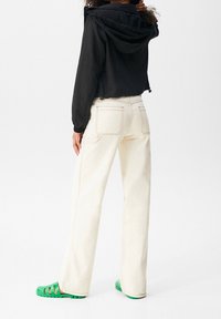 Veste noire courte à capuche, jean crème à jambes larges avec coutures visibles, et sandales en caoutchouc vertes avec semelle texturée.