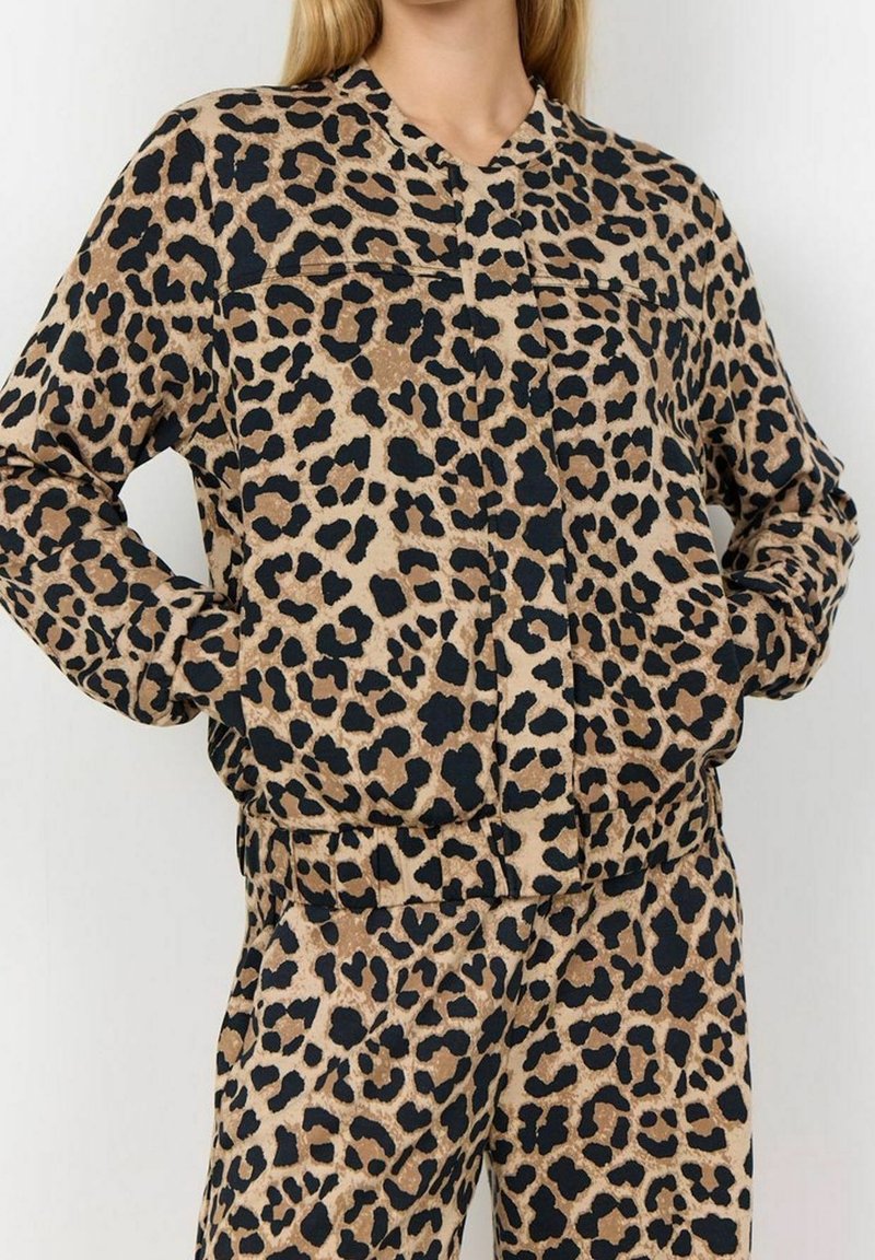 Femme portant une veste bomber et un pantalon assortis à imprimé léopard, mains dans les poches, debout devant un fond uni.