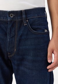 Jeans din denim de culoare bleu închis, cu cusături vizibile, un buzunar frontal și eticheta mărcii G-Star. Dispune de o închidere clasică cu buton și o textură netedă.