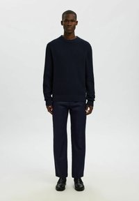 Uomo in piedi di fronte, indossa un maglione texturizzato blu scuro, pantaloni navy e scarpe eleganti nere su uno sfondo chiaro e semplice.