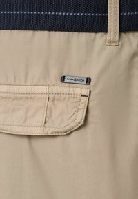 Beżowa kieszeń z tkaniny o zaokrąglonych krawędziach, metalowa metka z logo oraz teksturowana czarna górna część z poziomymi niebieskimi liniami jako akcentem.