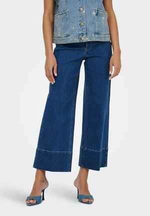 Nő derékig érő, bő szárú kék farmernadrágban, virágos hímzésű farmermellényben és kék, nyitott orrú magassarkú szandálban áll sima háttér előtt.