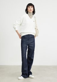 Witte v-hals trui, beige coltrui, donkerblauwe wijde jeans, witte sneakers en een lichtbeige schoudertas met een kettingstrap.