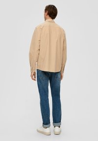 Beige Langarmbluse mit button-down Kragen, kombiniert mit blauen Jeans und weißen Sneakers; lässiger Schnitt, glatte Textur.