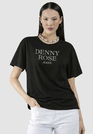 Donna con lunghi capelli neri indossa una maglietta nera con il logo "DENNY ROSE JEANS", pantaloni bianchi, una collana di perle e orecchini a cerchio, posando con la mano in tasca.