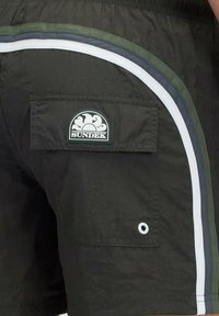 Pantaloni da bagno verde scuro con una tasca a patta e strisce bianche, blu e verdi lungo il lato. Presenta una toppa con logo sulla tasca.