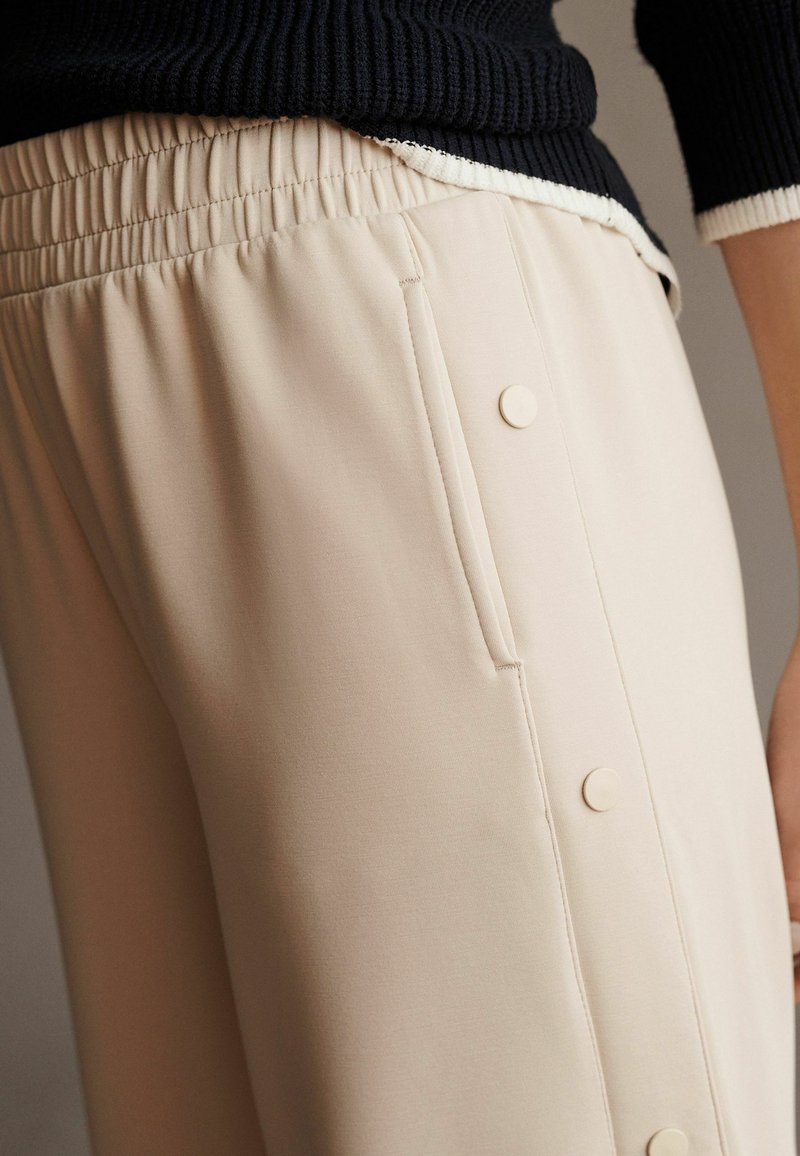 Beige bukser lavet af blødt materiale, med elastisk talje, sidelomme og dekorative knapper langs den ydre søm.