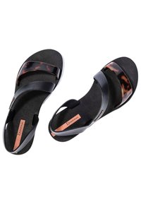 Sandalias negras con dos bandas de tiras brillantes y acentos de carey. Suela negra texturizada con etiqueta de marca en rosa. Diseño ligero y flexible.