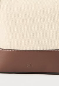 Beige strukturert stoffoverdel med en glatt brun lærbunn. Har kontraststing og en preget logo på den nedre kanten.