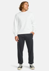 Witte crewneck sweatshirt met een gestructureerde afwerking, gecombineerd met zwarte joggerbroeken. De broek heeft een subtiele logo-detail en een relaxte pasvorm.