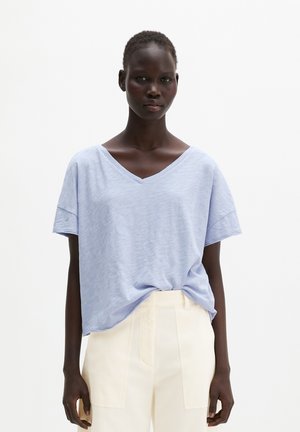 Vrouw met kort haar die een los lichtblauw T-shirt met V-hals draagt, ingestopt in een hooggetailleerde crème broek, staand tegen een effen witte achtergrond.