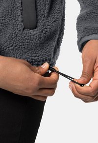 Pullover in pile grigio con superficie testurizzata, che mostra mani che regolano un cordino nero all'orlo, posto su uno sfondo bianco.
