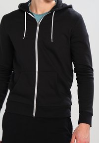 Sudadera con cremallera negra hecha de una tela suave. Cuenta con capucha ajustable, bolsillos frontales y detalles en cremallera blanca. Textura suave.