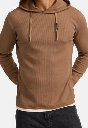 Mand iført brun hættejakke i vaffelstrik med snore og mørke bukser, hænderne bag ryggen, på en ensfarvet hvid baggrund.