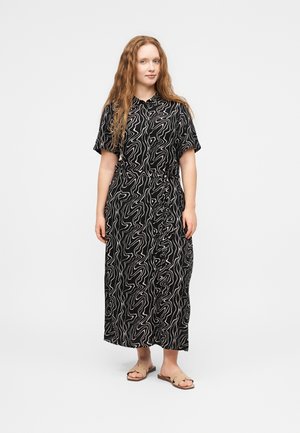 Kvinde med langt bølget hår iført en sort-hvid abstrakt mønstret midi-kjole og beige slip-on sandaler på en hvid baggrund.