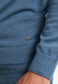 Suéter de punto azul con puños y dobladillo acanalados, y una pequeña etiqueta ovalada de plata en la parte inferior frontal. Textura suave y diseño casual.