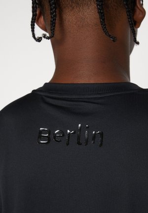 Gros plan sur le haut du dos d'une personne aux cheveux tressés, portant un t-shirt noir avec un texte brillant "Berlin" dans le dos près de l'encolure.