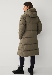 Frau in einem knielangen, olivgrünen, gepolsterten Wintermantel mit Kapuze, schwarzen Leggings und schwarzen Knöchelstiefeln, die an einer weißen Wand steht.