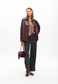 Giacca di pelle bordeaux lucida sopra una blusa con stampa leopardata, abbinata a jeans grigi a gamba larga e tacchi a punta bordeaux. Piccola borsa in pelliccia.