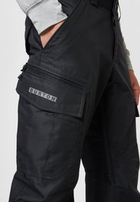 Zwarte cargobroek met een gestructureerde afwerking, voorzien van meerdere zakken, een ritssluiting en een grijs "BURTON" logo aan de zijkant.