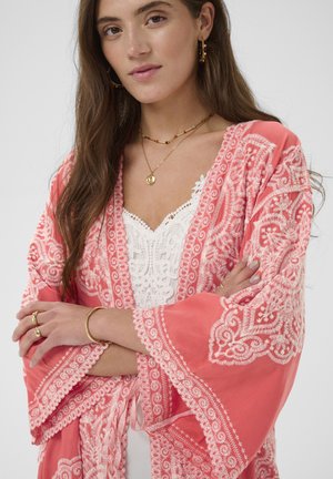 Jonge vrouw met lang bruin haar, gekleed in een koraalroze geborduurde kimono over een wit kanten topje, geaccentueerd met gouden sieraden en met gekruiste armen.