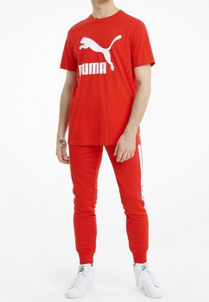 Homme portant un t-shirt Puma rouge avec logo et un pantalon rouge assorti à rayures blanches, associé à des baskets blanches.