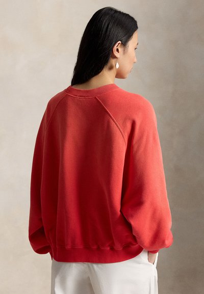 Γυναίκα με σκούρα μαλλιά που φοράει κόκκινο φούτερ με στρογγυλή λαιμόκοψη και λευκό παντελόνι, στέκεται πλαγιαστά με τα χέρια στις τσέπες μπροστά σε ουδέτερο φόντο.