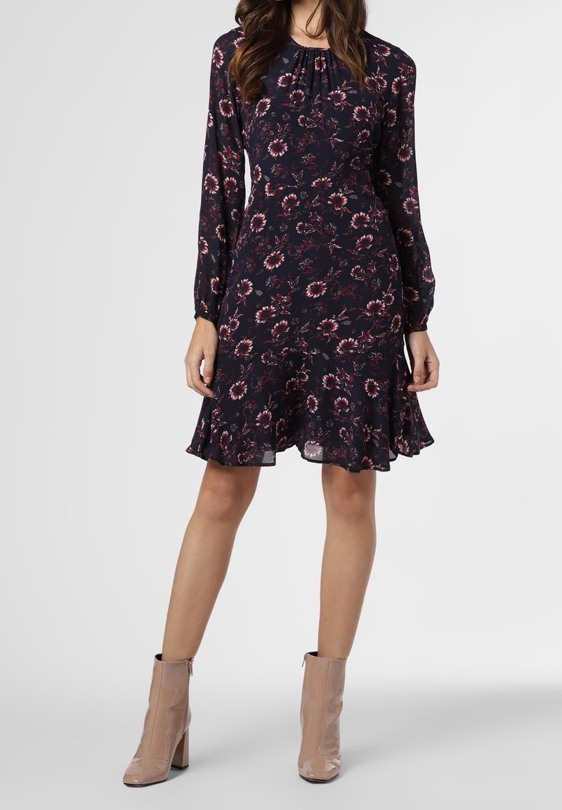 Femme portant une robe noire à manches longues à motif floral avec un ourlet évasé et des bottines beiges, debout devant un fond uni.