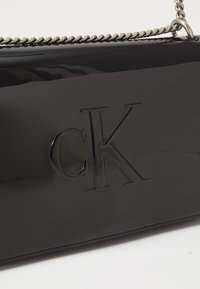Black patent leather handbag featuring embossed "CK" logo, chain-accented strap, and smooth, shiny texture. Minimalist rectangular design.