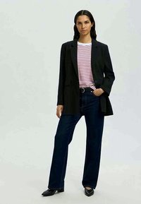 Blazer preto com lapelas recortadas, usado sobre uma camisa listrada de branco e vermelho, combinado com jeans de perna larga azul escuro e sapatos pretos de bico fino.