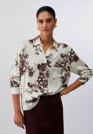 Donna con capelli scuri che indossa orecchini a cerchio dorati, una camicetta crema con bottoni e stampa floreale marrone, e una gonna scura, posa con una mano sul fianco.