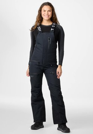 Młoda kobieta o długich kręconych włosach, ubrana w czarne narciarskie spodnie ogrodniczki oraz czarną bluzkę z długim rękawem, stoi i uśmiecha się na białym tle.