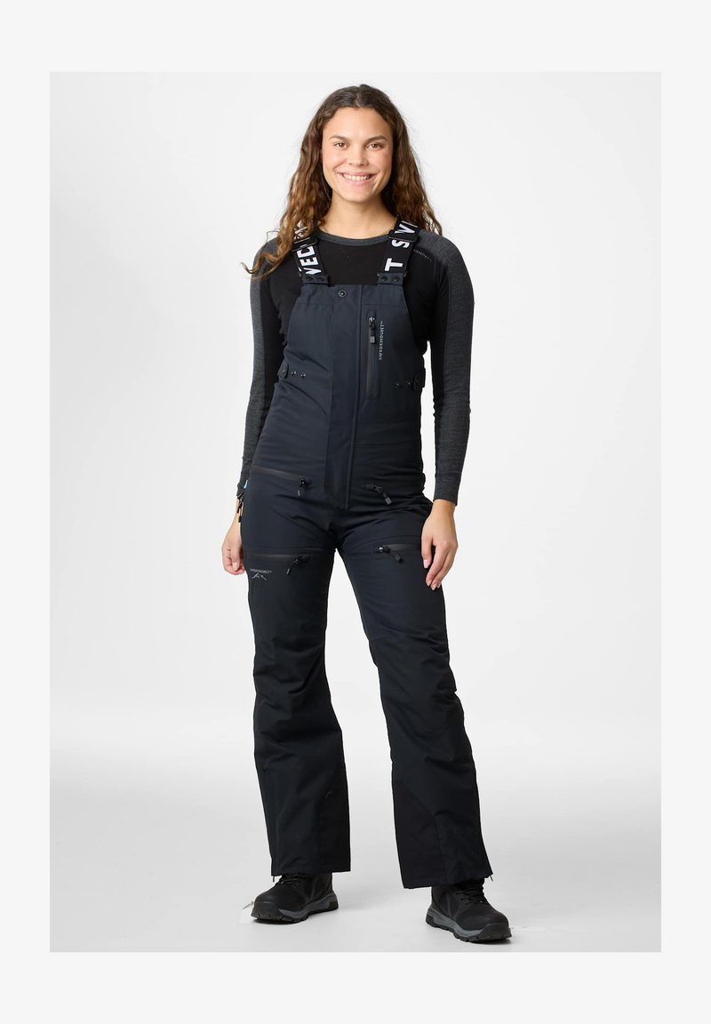 Giovane donna con lunghi capelli ricci che indossa pantaloni da sci neri con bretelle e una maglia nera a maniche lunghe, in piedi e sorridente su sfondo bianco.