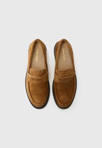 Mocassins en suède marron avec un bout arrondi et une sangle décorative, sur un fond clair.