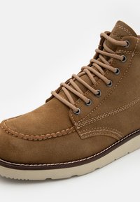 Zapatos altos de ante marrón con cordones beige, ojales de metal y detalles de costura. La suela es de color crema y tiene textura para ofrecer un buen agarre.