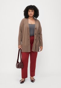 Brown knit cardigan with button closure, gray tank top, red trousers, and dark red bag. Flat shoes with chain detail. Textured fabric.