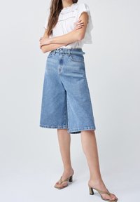 Culottes en denim bleu clair avec une coupe ample, taille haute et ourlet brut. Associés à un chemisier blanc et des sandales à talons vertes.