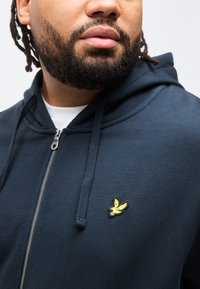 Άνδρας με γενειάδα που φοράει μπλε σκουρόχρωμο φούτερ με φερμουάρ και κέντημα ενός κίτρινου πουλιού στο στήθος και λευκό πουκάμισο από κάτω, στραμμένος ελαφρώς προς τα αριστερά.