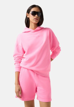 Vrouw draagt een felroze hoodie en bijpassende short met donkere oversized zonnebril, poseert met één hand op haar heup tegen een lichte achtergrond.