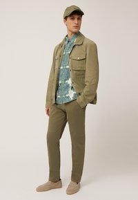 Olive green suede jacket with chest pockets, vibrant patterned shirt, olive trousers, and beige slip-on shoes. Matched with a cap.