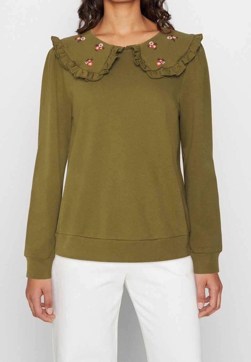 Haut à manches longues vert olive avec un grand col à volants présentant de petites fleurs brodées roses et rouges, associé à un pantalon blanc.