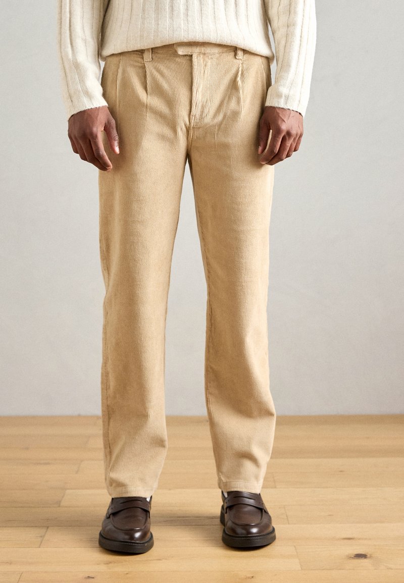 Pantaloni in corduroy beige con taglio dritto, caratterizzati da una superficie testurizzata e pieghe frontali, abbinati a scarpe marroni scure.