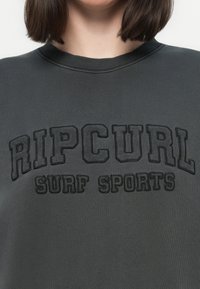 Persona che indossa un felpa grigio scuro con testo nero in rilievo che recita "RIP CURL SURF SPORTS" sul petto.