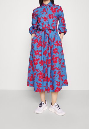 Frau trägt ein blaues Midikleid mit rotem Blumenmuster, tailliert mit Gürtel, dreiviertellangen Ärmeln und weißen Sneakers mit schwarzen Schnürsenkeln, Hände in den Taschen.