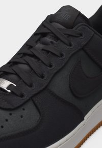 Κοντινό πλάνο ενός μαύρου παπουτσιού Nike Air Force 1 που δείχνει τα κορδόνια, την περφορεμένη περιοχή στα δάχτυλα και το ανάγλυφο λογότυπο Nike στην άσπρη σόλα με καφέ εξωτερική σόλα.