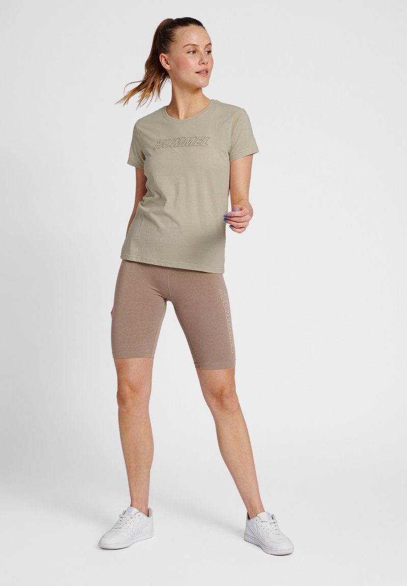 Hellgrünes T-Shirt mit geprägtem Logo, kombiniert mit beigefarbenen Biker-Shorts, die ein seitliches Logo haben. Die Schuhe sind weiße Sportschuhe.