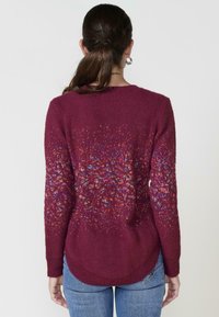 Burgundi színű pulóver, texturált felülettel, színes foltos mintákkal, hosszú ujjakkal, és lekerekített hátsó heméllyel.