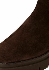 Gros plan d'une botte en daim brun foncé avec des coutures détaillées et une semelle en caoutchouc épaisse et rainurée.