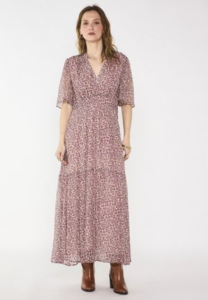 Donna che indossa un lungo vestito floreale viola con maniche corte e stivaletti marroni, in piedi davanti a uno sfondo bianco uniforme.